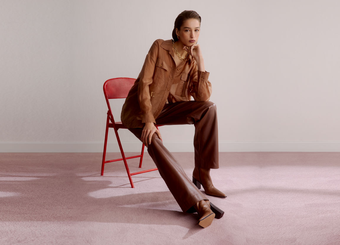 Modelo sentada numa cadeira usando look todo marrom com calça de couro, camisa e bota.